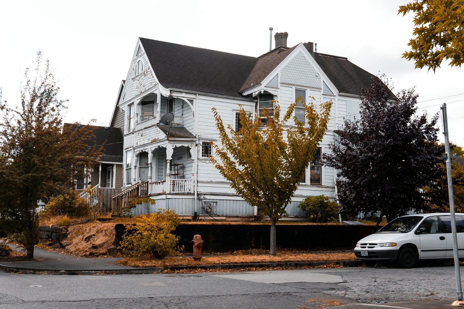 A weathered white wooden house with autumn trees, parked car, and fall ambiance on a suburban street.